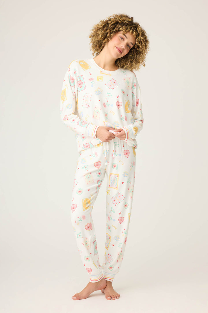 A curly-haired person stands barefoot in the P.J. Salvage Pant Set LUCKY PJ SET LUCKY ME, a white pajama set with colorful playing card and heart patterns, against a plain light background.