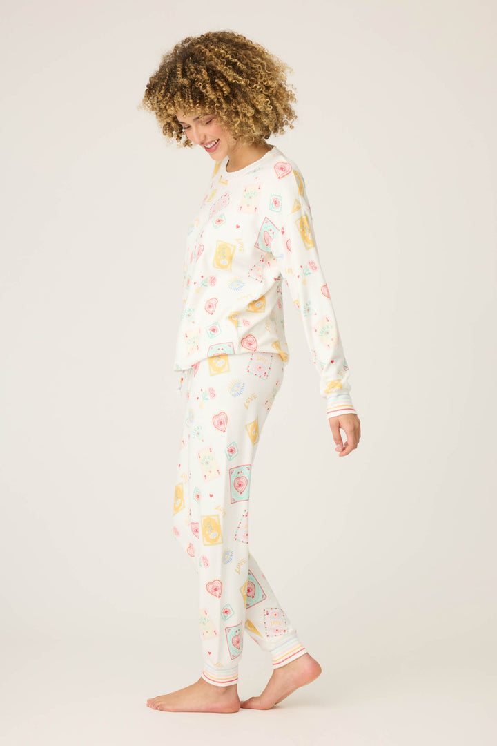 A person with curly hair stands barefoot against a plain background, wearing the P.J. Salvage Pant Set LUCKY PJ SET LUCKY ME—a matching long-sleeve pajama set with a colorful food-themed print—looking down and smiling.