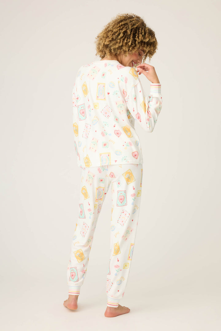 A person with curly hair stands barefoot, facing away from the camera, wearing the P.J. Salvage Pant Set LUCKY PJ SET LUCKY ME—matching white pajamas with a colorful playing card and heart pattern on both the top and pants.