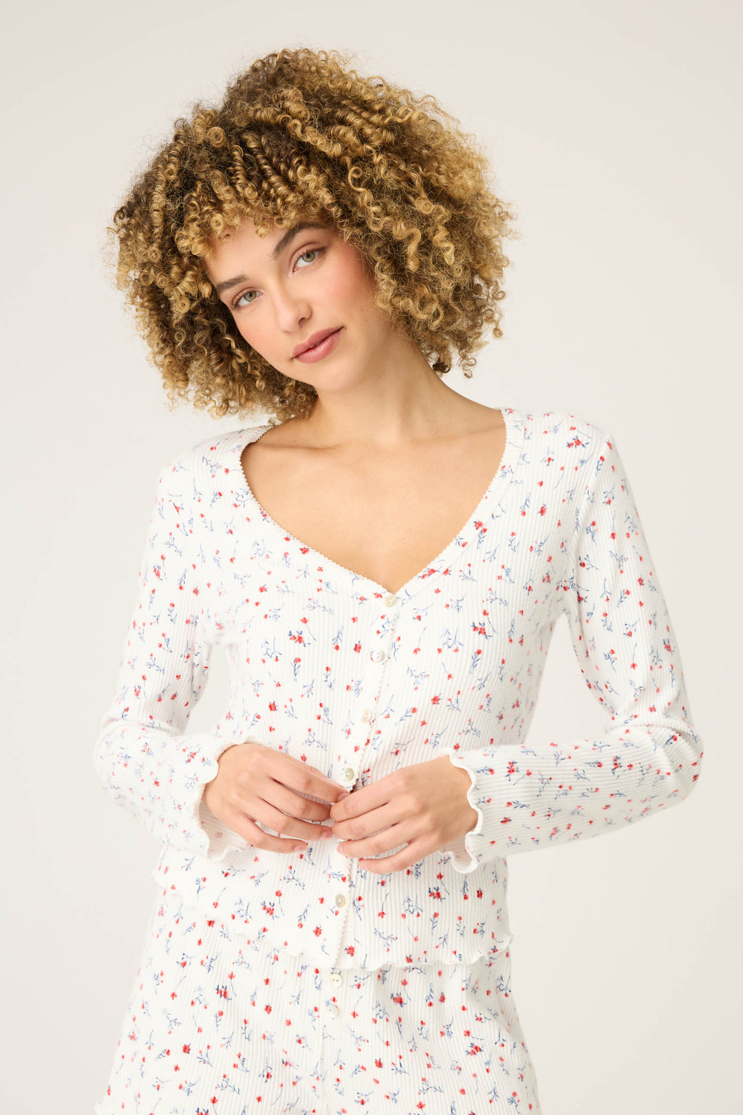 A woman with curly blonde hair models the P.J. Salvage ROSE CARDIGAN LA ROSE, standing against a plain light background and looking slightly to the side with her hands together.