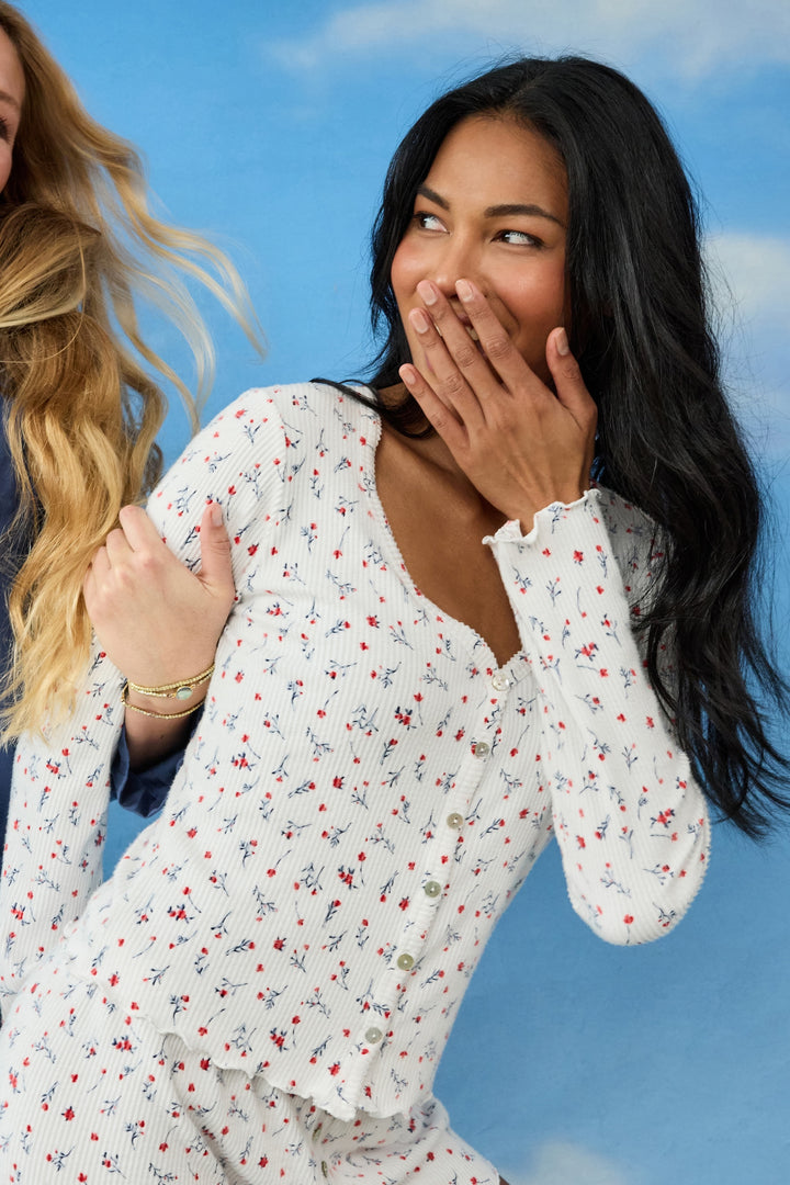 A woman with long black hair smiles with her hand over her mouth, wearing the P.J. Salvage ROSE CARDIGAN LA ROSE, standing against a blue sky with clouds. Another person with long blonde hair is partly visible beside her.