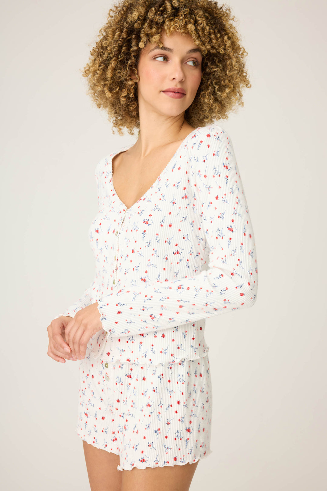 A person with curly hair wears the P.J. Salvage ROSE CARDIGAN LA ROSE, a long-sleeve white floral top with matching shorts, standing against a plain light background and looking slightly to the side.