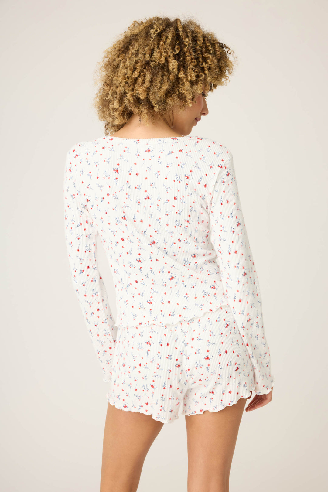 A person with curly hair faces away from the camera, wearing the ROSE CARDIGAN LA ROSE by P.J. Salvage—a white ribbed set with small red and blue floral prints and scalloped edges.