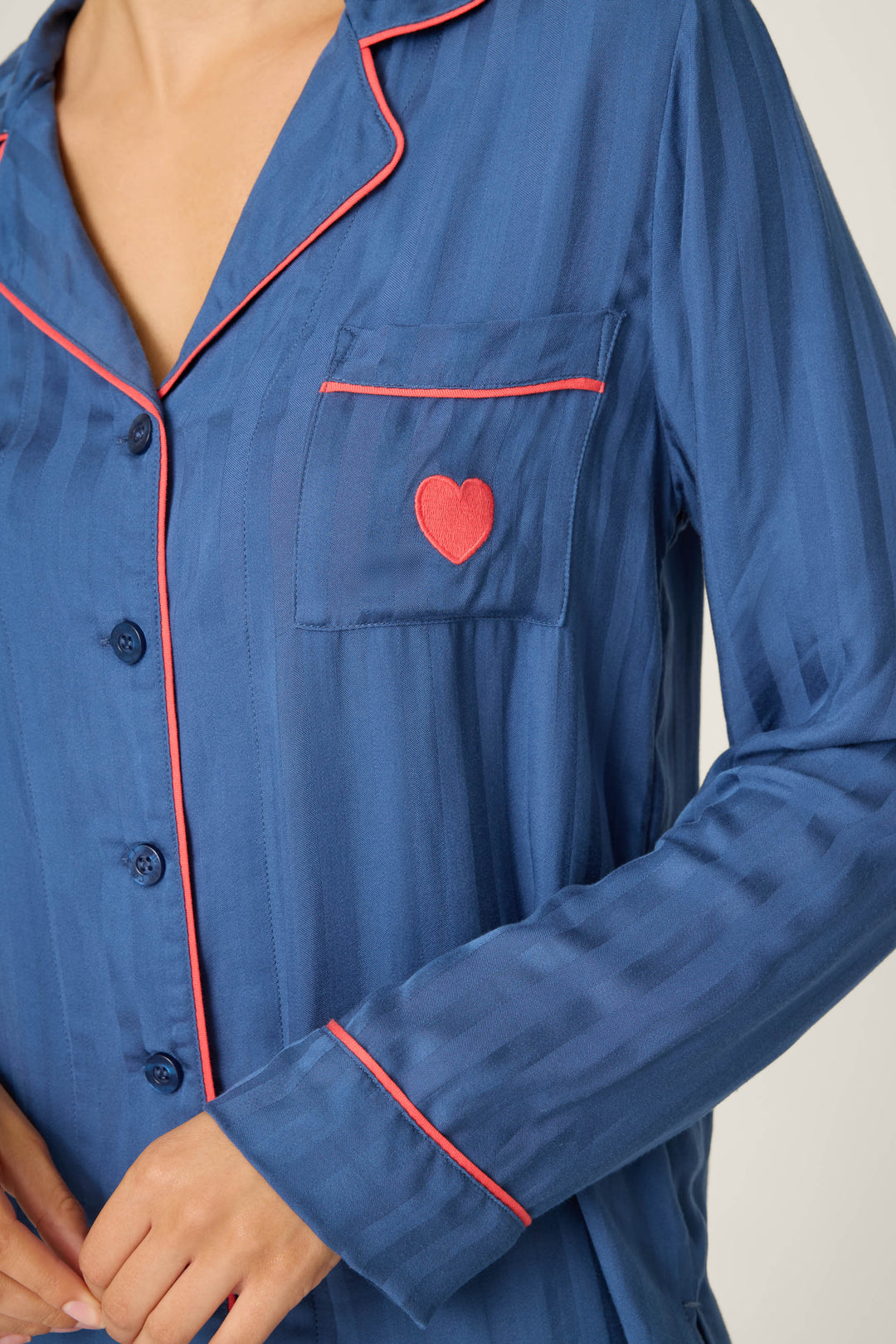 A person wears the P.J. Salvage La Rose Striped Long Sleeve Top, a blue jacquard sateen pajama top with red piping, buttons, and a chest pocket embroidered with a heart. Their hands are partially visible at the bottom.