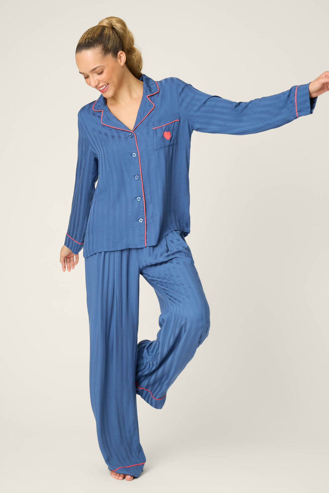 A person wears the P.J. Salvage La Rose Striped Long Sleeve Top, standing on one foot with a smile and looking down. The top has a heart-embroidered pocket and pink trim. The background is plain and light-colored.
