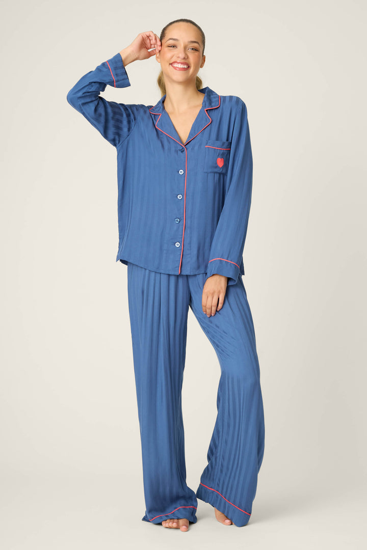 A woman stands barefoot, smiling, wearing the P.J. Salvage STRIPES PJ SET LA ROSE—a blue pajama set with pink piping, button-up shirt with chest pocket, and loose-fitting pants. She raises one hand to touch her head.