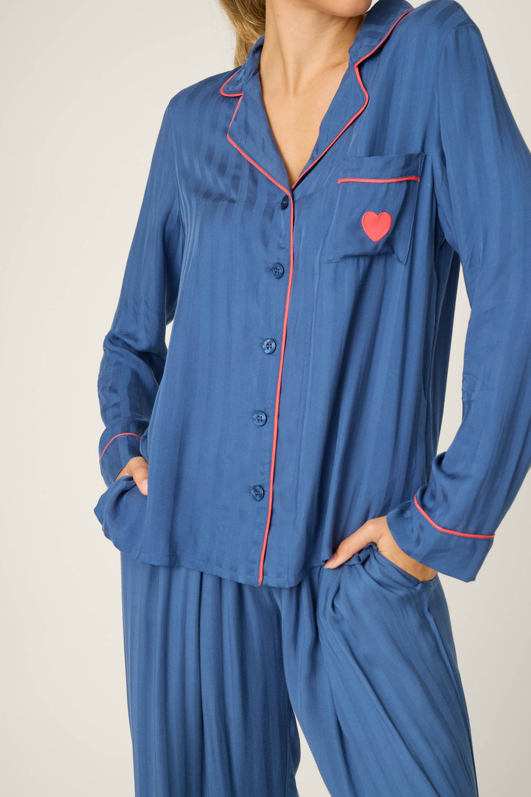A person wears the STRIPES PJ SET LA ROSE by P.J. Salvage—a blue, long-sleeve button-up pajama top with red piping and a small red heart on the chest pocket, paired with matching loose-fitting pants.