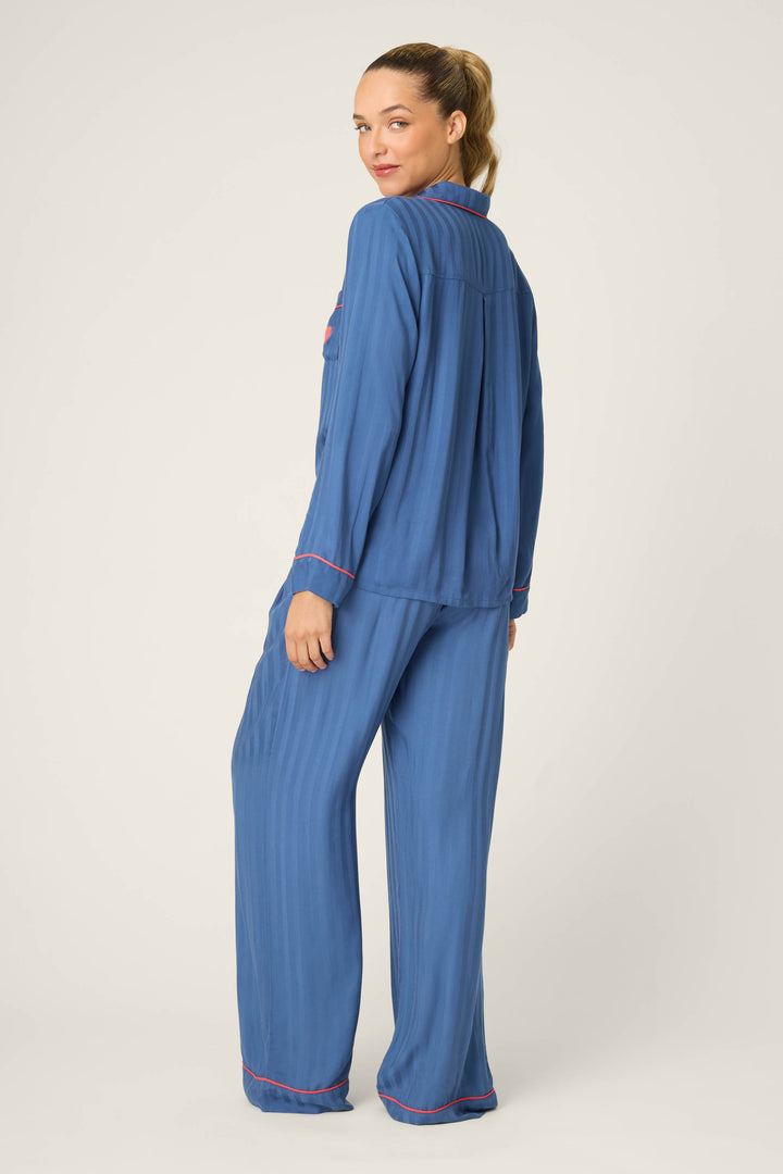 A woman faces away and looks over her shoulder, wearing the P.J. Salvage STRIPES PJ SET LA ROSE. The loose-fitting pajama set features blue fabric with pink trim, set against a plain light-colored background.