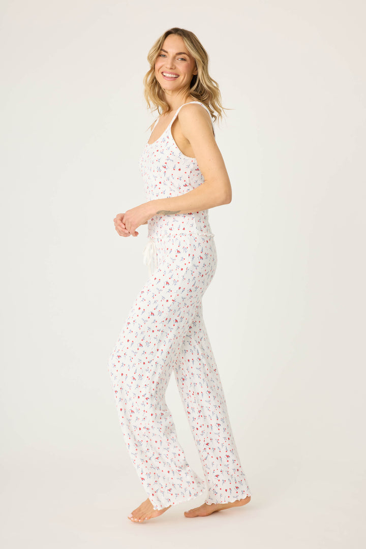 A woman stands barefoot on a white background, smiling and looking to the side, wearing the P.J. Salvage FLORAL PJ SET LA ROSE—a white sleeveless pajama set with a small red and blue floral print and drawstring waist.