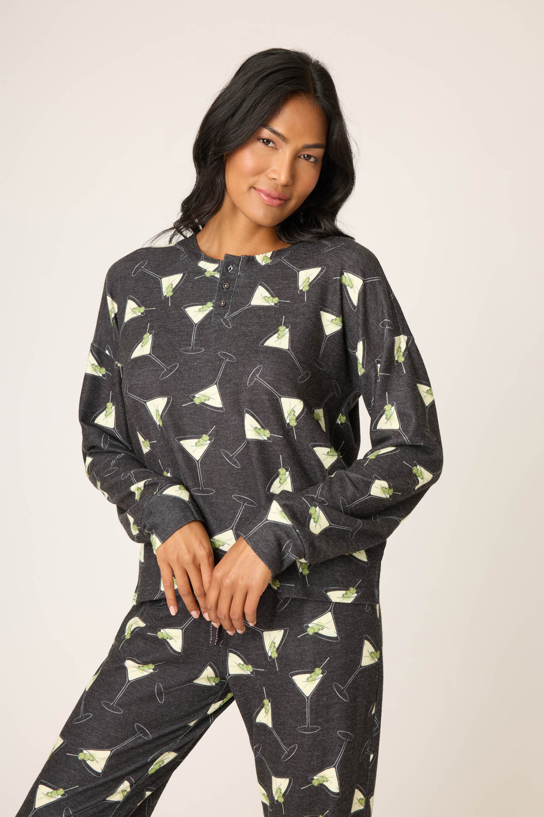 A person with long, dark hair wears the P.J. Salvage Merrier With A Martini Long Sleeve Top—a dark grey pajama top with a martini glass print and buttons at the neckline—with matching pants, standing against a plain background.