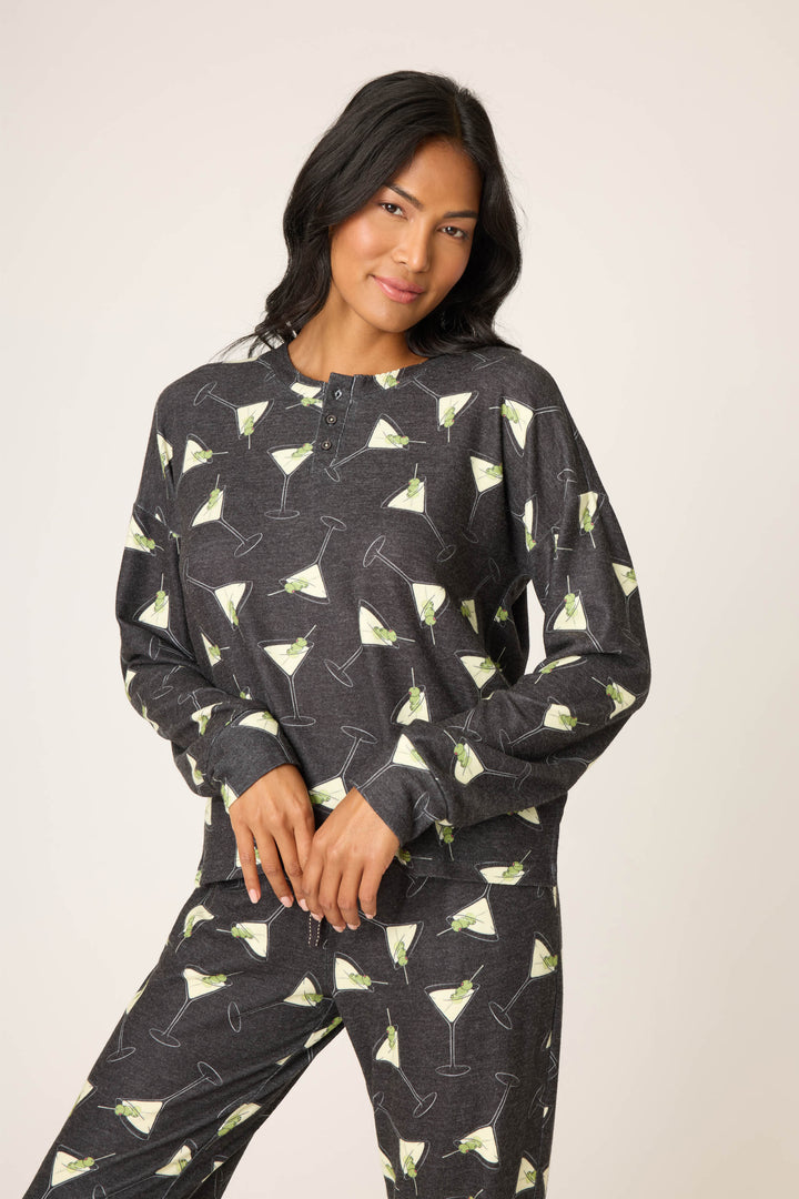 A person with long, dark hair wears the P.J. Salvage Merrier With A Martini Long Sleeve Top—a dark grey pajama top with a martini glass print and buttons at the neckline—with matching pants, standing against a plain background.