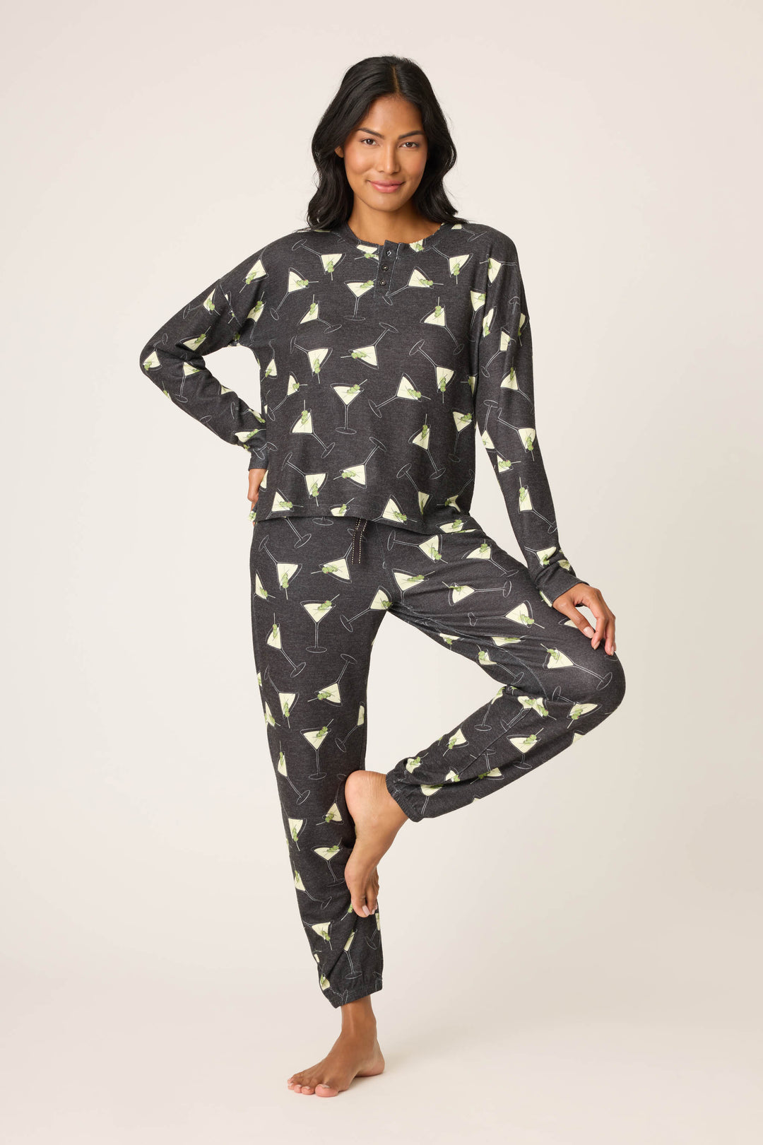 A person stands barefoot on one leg wearing the P.J. Salvage "Merrier With A Martini" Long Sleeve Top, featuring a martini glass pattern, against a plain light background.