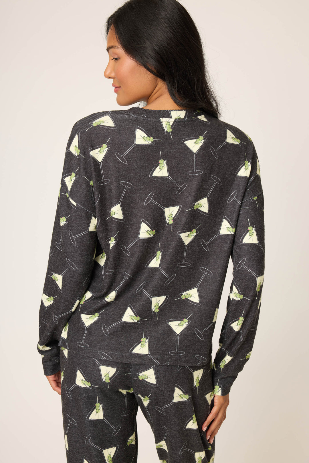 A woman with long dark hair models the P.J. Salvage "Merrier With A Martini" Long Sleeve Top, shown from the back with her head turned slightly, featuring a matching dark gray pajama set with an all-over martini glass print.