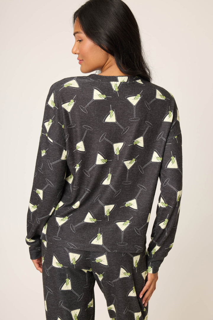 A woman with long dark hair models the P.J. Salvage "Merrier With A Martini" Long Sleeve Top, shown from the back with her head turned slightly, featuring a matching dark gray pajama set with an all-over martini glass print.