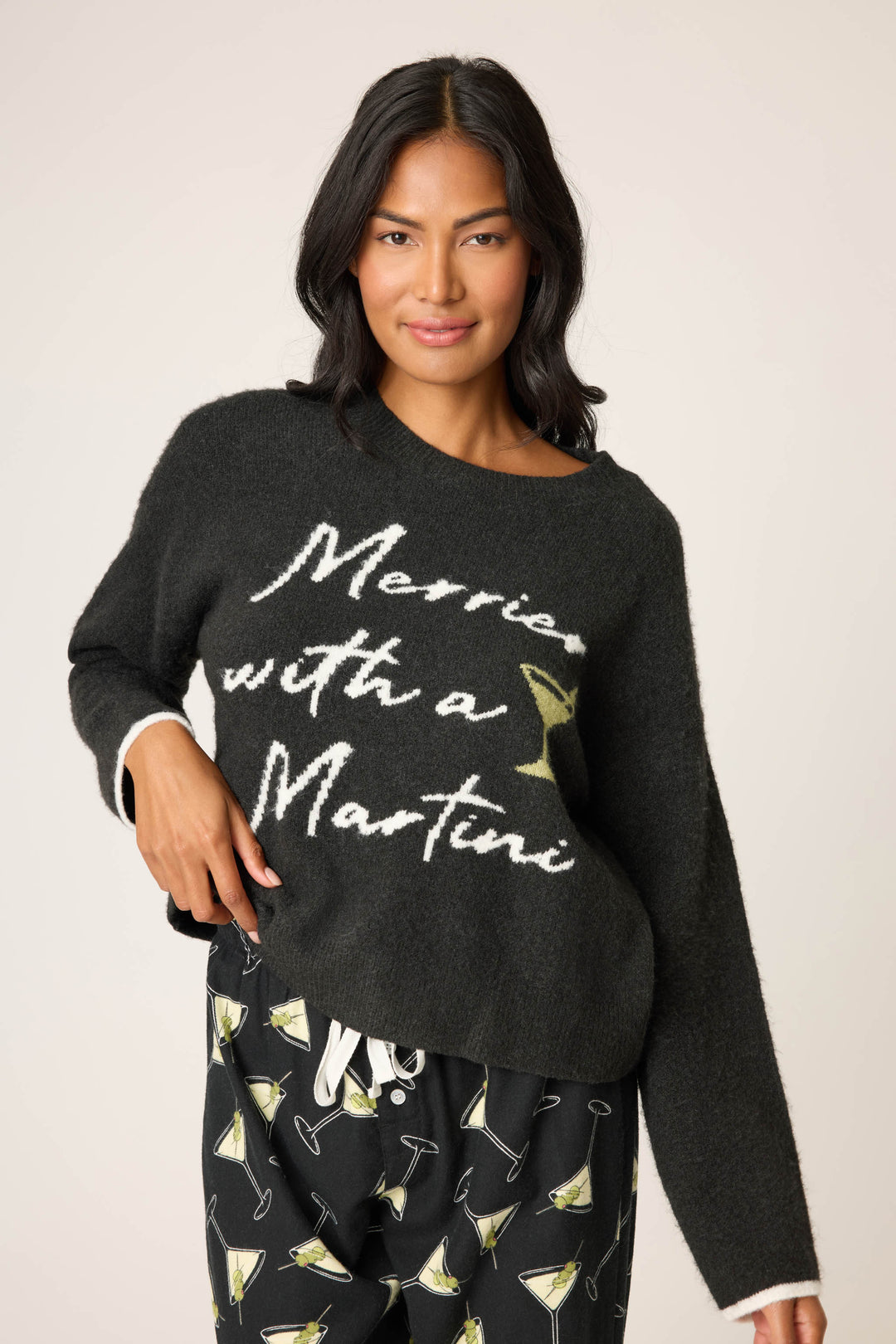 A woman with medium-length dark hair wears the P.J. Salvage Merrier With A Martini Snuggle Sweater, featuring the phrase "Merrier with a Martini" and martini glass graphic, paired with black pants decorated with martini glasses.