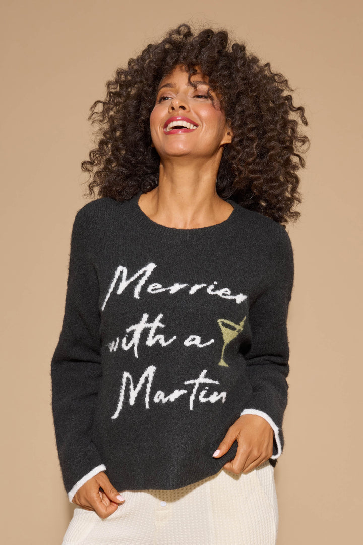 A woman with curly hair smiles and looks to the side while wearing the P.J. Salvage Merrier With A Martini Snuggle Sweater, a dark top featuring "Merrier with a Martini" in white script and a small martini glass graphic.
