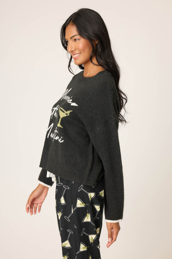 A woman with long dark hair smiles in profile, wearing the P.J. Salvage Merrier With A Martini Snuggle Sweater, featuring writing and a martini glass graphic, paired with black pants adorned with martini glass prints.
