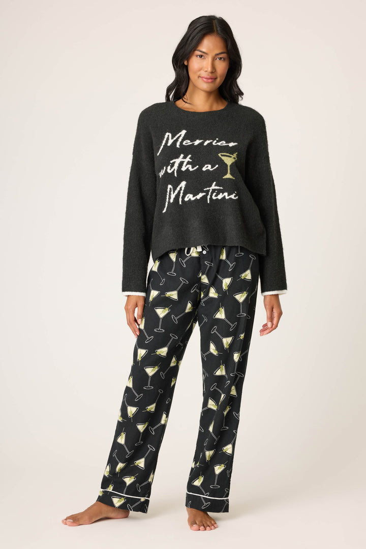 A woman stands barefoot, wearing P.J. Salvage’s Merrier With A Martini Snuggle Sweater and matching black pajama pants with martini glass prints, facing forward against a plain background.