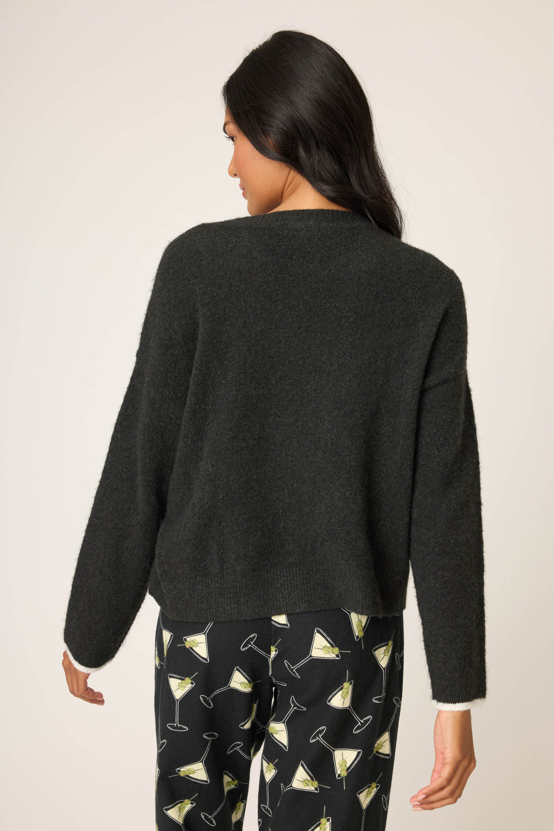 A woman with long dark hair is seen from the back, wearing the P.J. Salvage Merrier With A Martini Snuggle Sweater and matching martini glass print pants, standing against a plain light background.