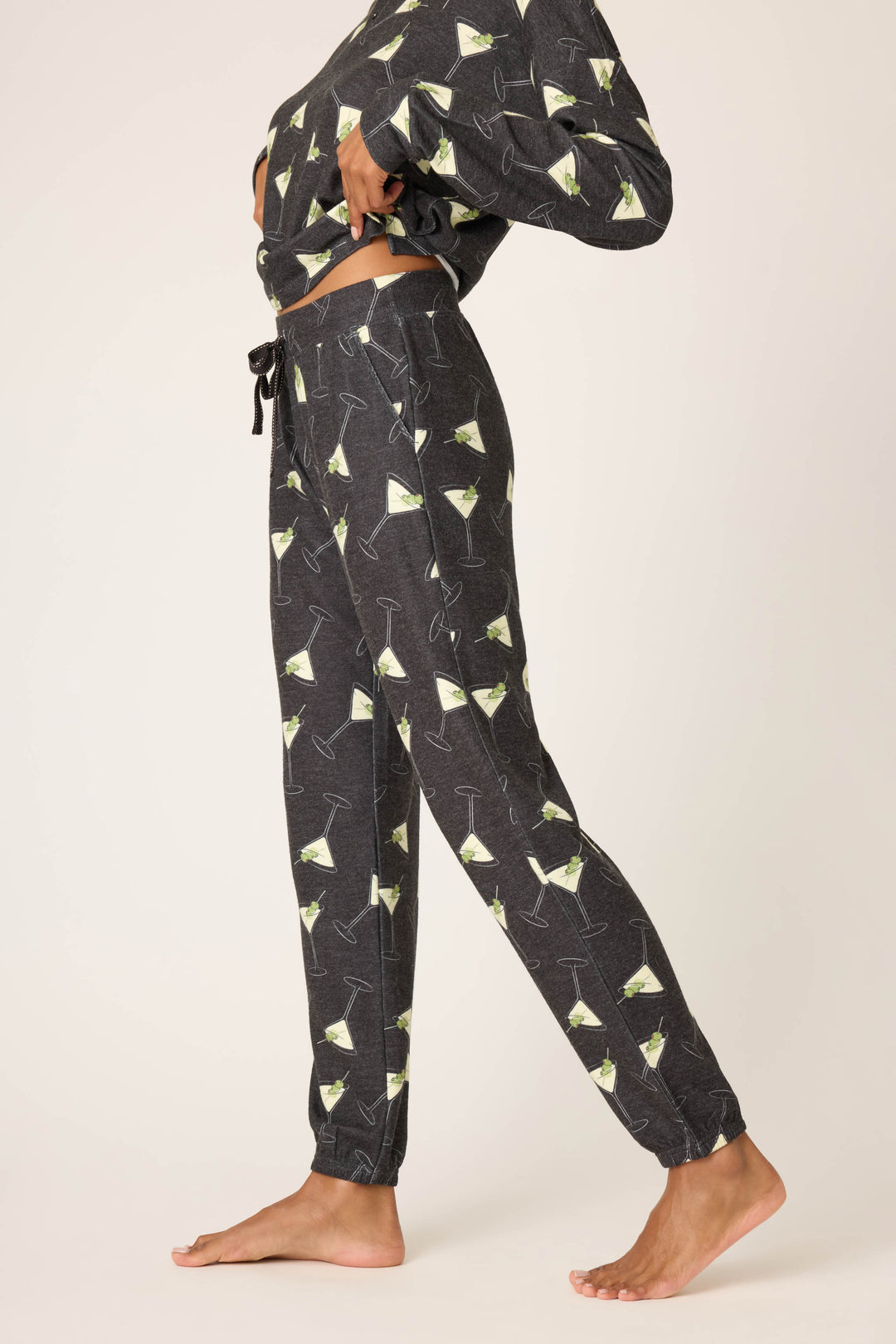 A person stands barefoot in profile wearing P.J. Salvage’s Merrier With A Martini Banded Pant and matching top with a martini glass print, hand on hip, against a plain light background.