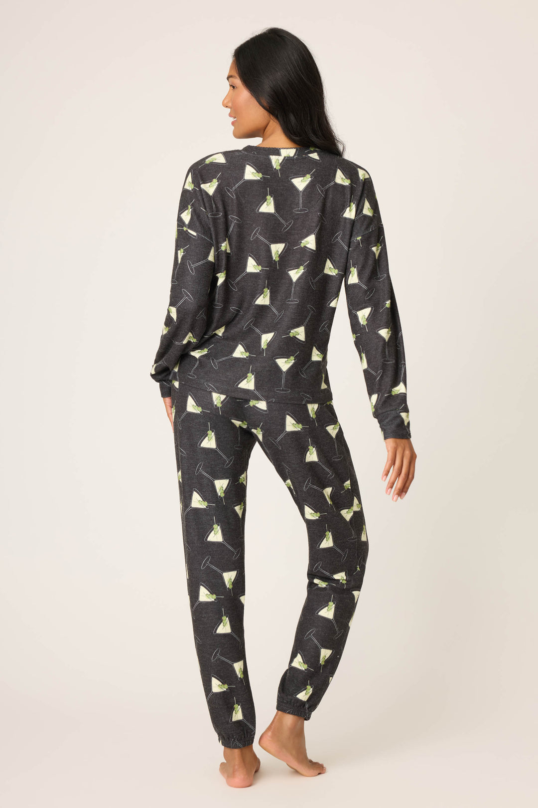 A woman with dark hair stands barefoot facing away, wearing the P.J. Salvage Merrier With A Martini PJ Set—a matching long-sleeve top and pants featuring a martini glass print on a dark background, set against a plain light backdrop.