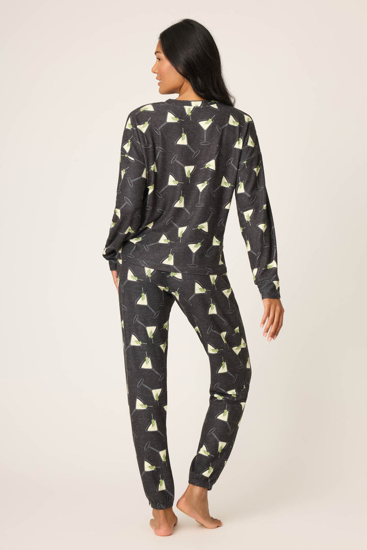 A woman with dark hair stands barefoot facing away, wearing the P.J. Salvage Merrier With A Martini PJ Set—a matching long-sleeve top and pants featuring a martini glass print on a dark background, set against a plain light backdrop.