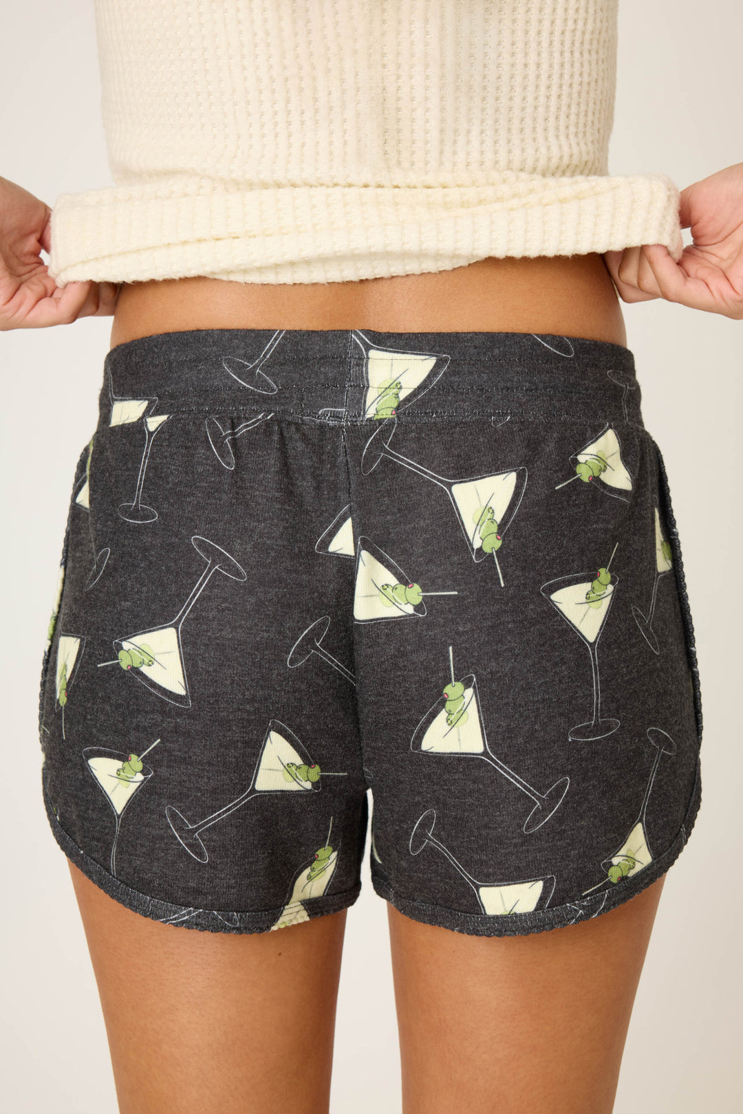 A person stands with their back to the camera, pulling up a cream ribbed sweater to reveal the waistband of the P.J. Salvage Merrier With A Martini Short, featuring a dark gray fabric with a martini glass and olive print.