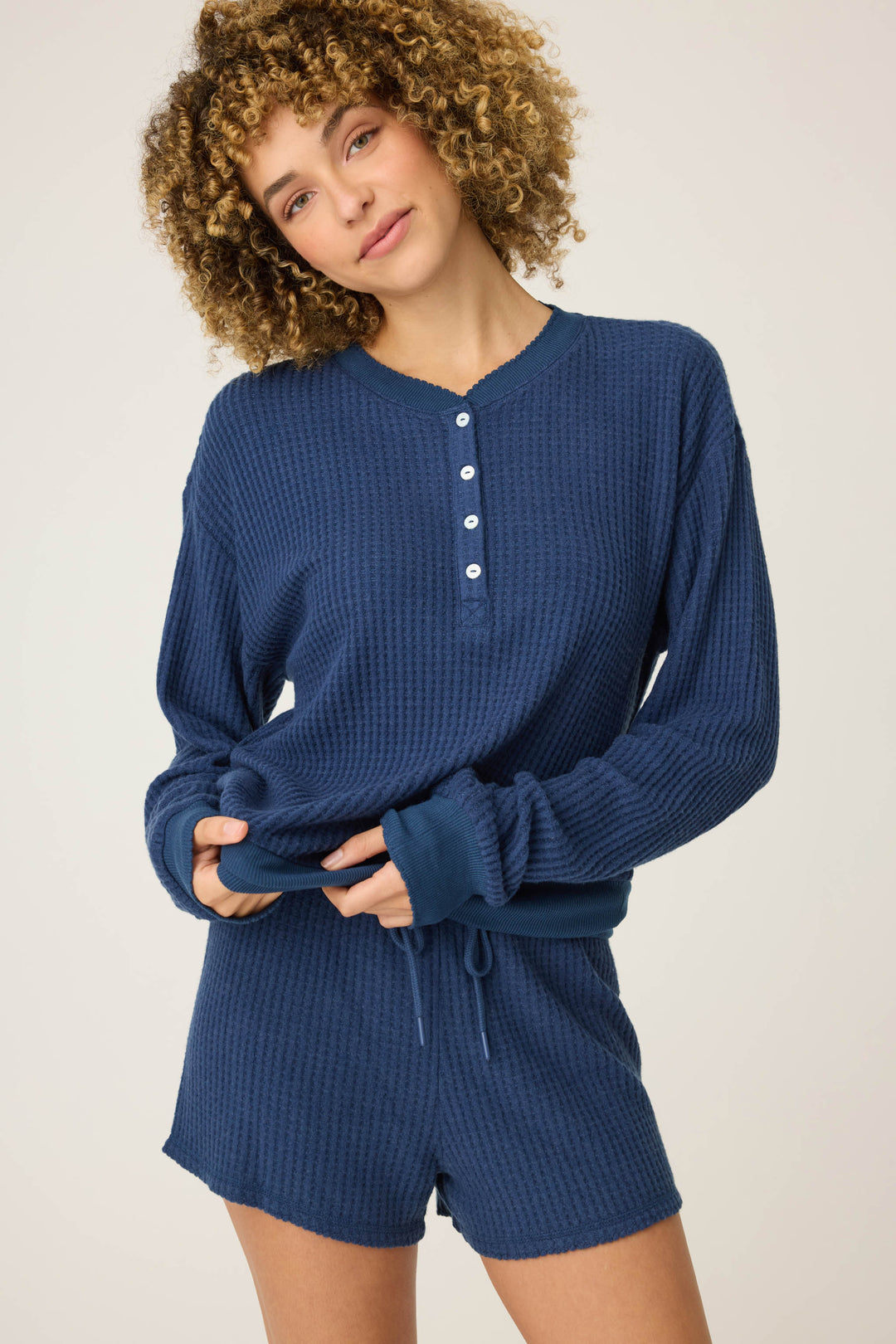 A person with curly hair wears the P.J. Salvage Morning Waffles Solid Navy Long Sleeve Top and matching shorts, standing against a plain light background, holding the shirt at the waist with a neutral expression.
