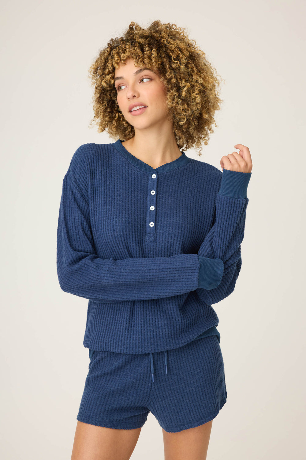 A person with curly hair wears the P.J. Salvage Morning Waffles Solid Navy Long Sleeve Top, standing against a plain light background and looking slightly to the side.