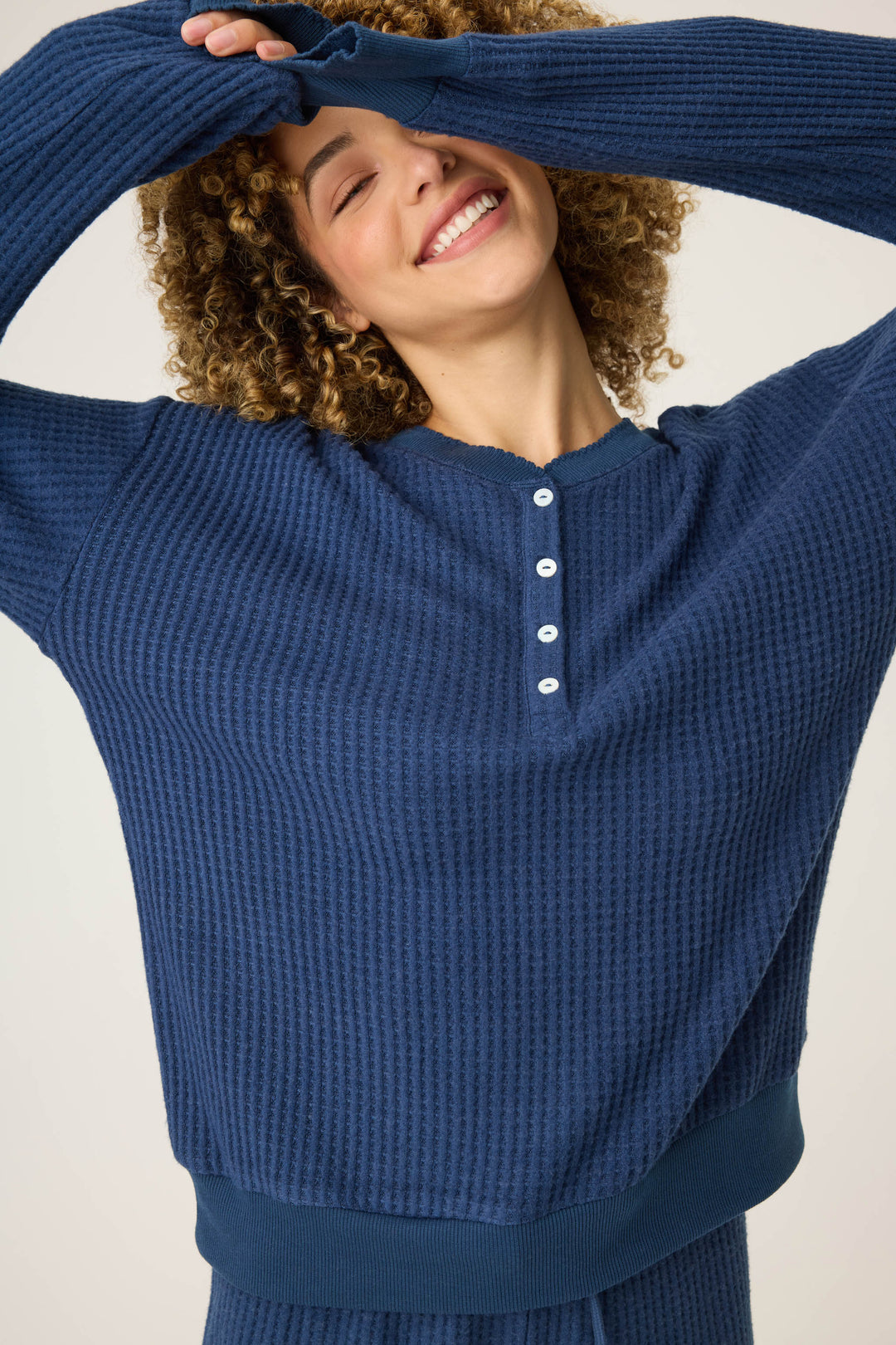 A person with curly hair smiles and raises their arms, wearing the P.J. Salvage Morning Waffles Solid Navy Long Sleeve Top. The shirt is textured with four front buttons, and the background is plain and light-colored.