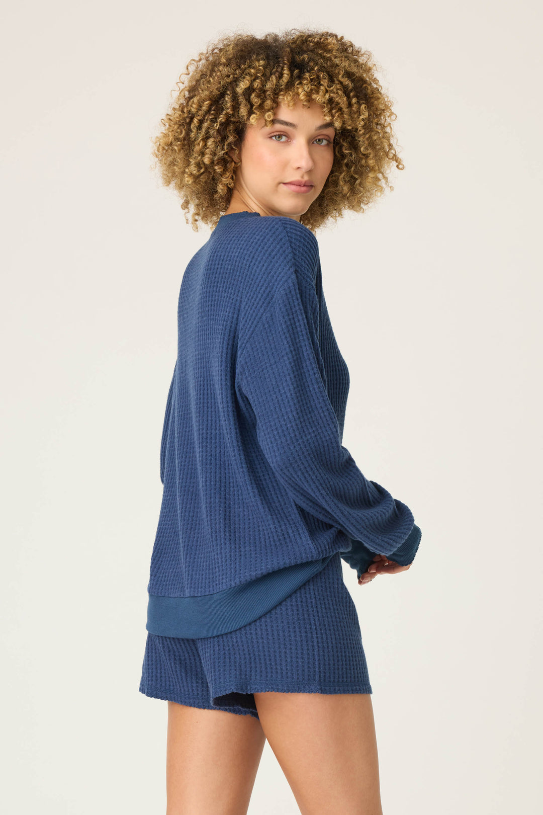 A person with curly hair wears the P.J. Salvage Morning Waffles Solid Navy Long Sleeve Top, paired with matching navy shorts, standing against a plain light background and glancing back at the camera.