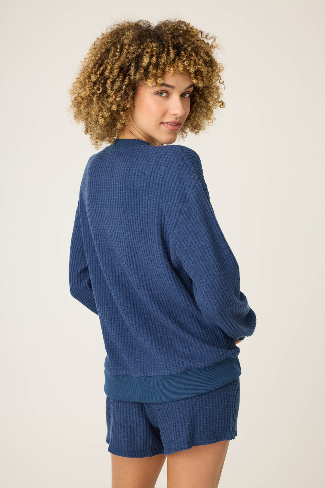 A person with curly hair wearing the P.J. Salvage Morning Waffles Solid Navy Long Sleeve Top stands turned slightly to the side, looking over their shoulder against a plain light background.