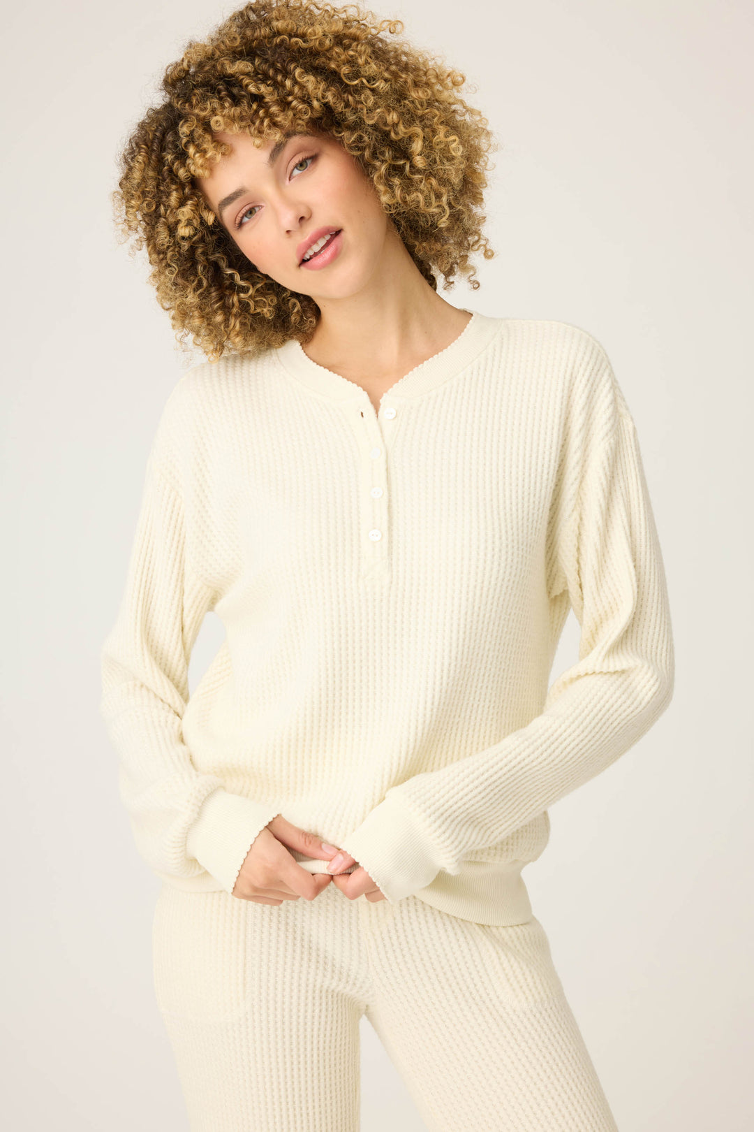 A person with curly hair wears the P.J. Salvage Morning Waffles Solid Ivory Long Sleeve Top, standing with hands clasped in front of a plain light background.