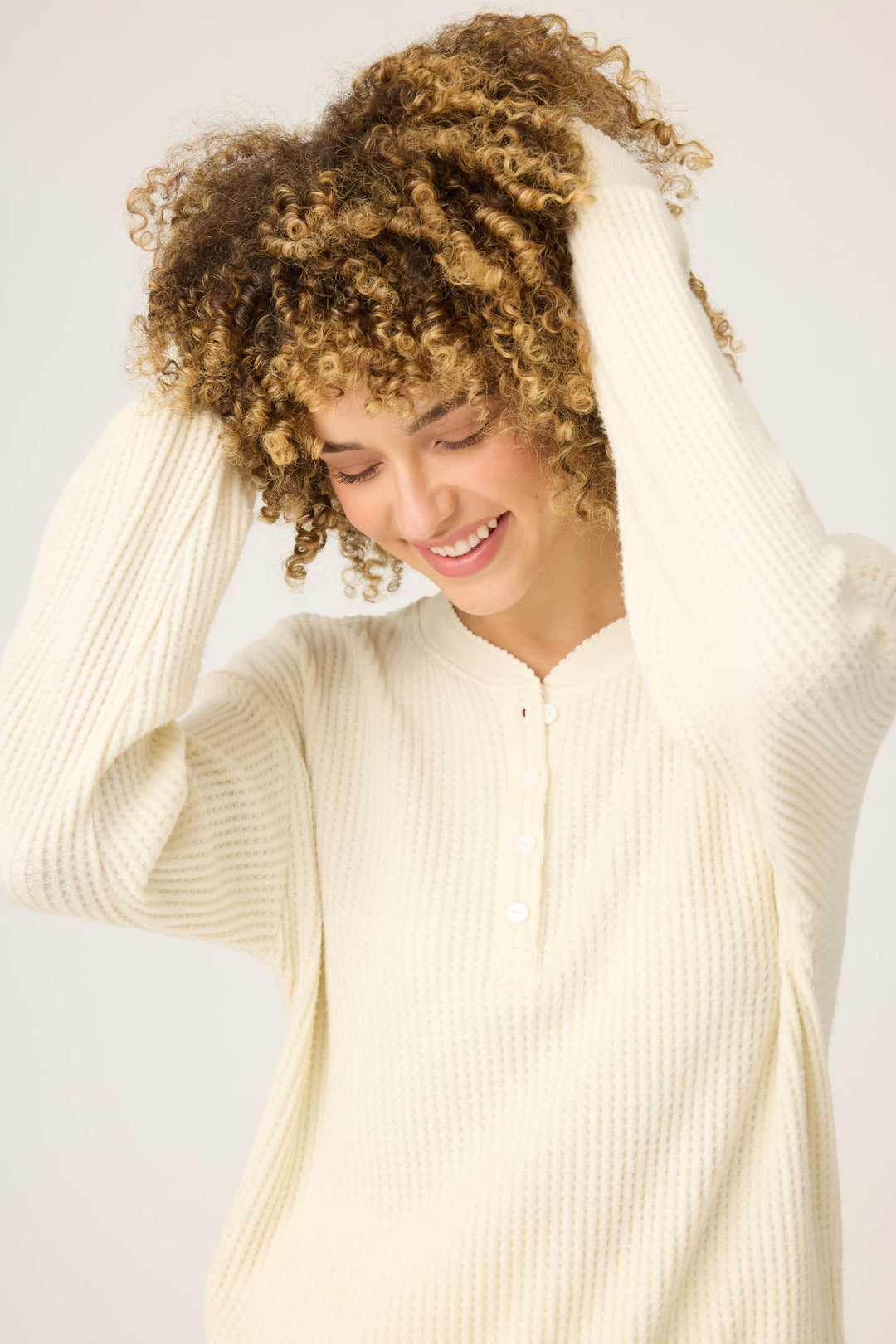 A person wearing the P.J. Salvage Morning Waffles Solid Ivory Long Sleeve Top smiles with eyes closed, lifting their curly hair with both hands against a plain light background.