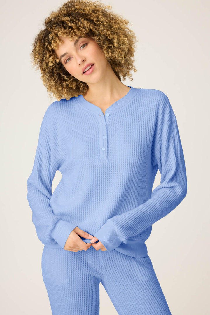 A person with curly hair wears the P.J. Salvage Morning Waffles Solid Peri Long Sleeve Top, standing against a plain light background and looking at the camera with a relaxed expression.