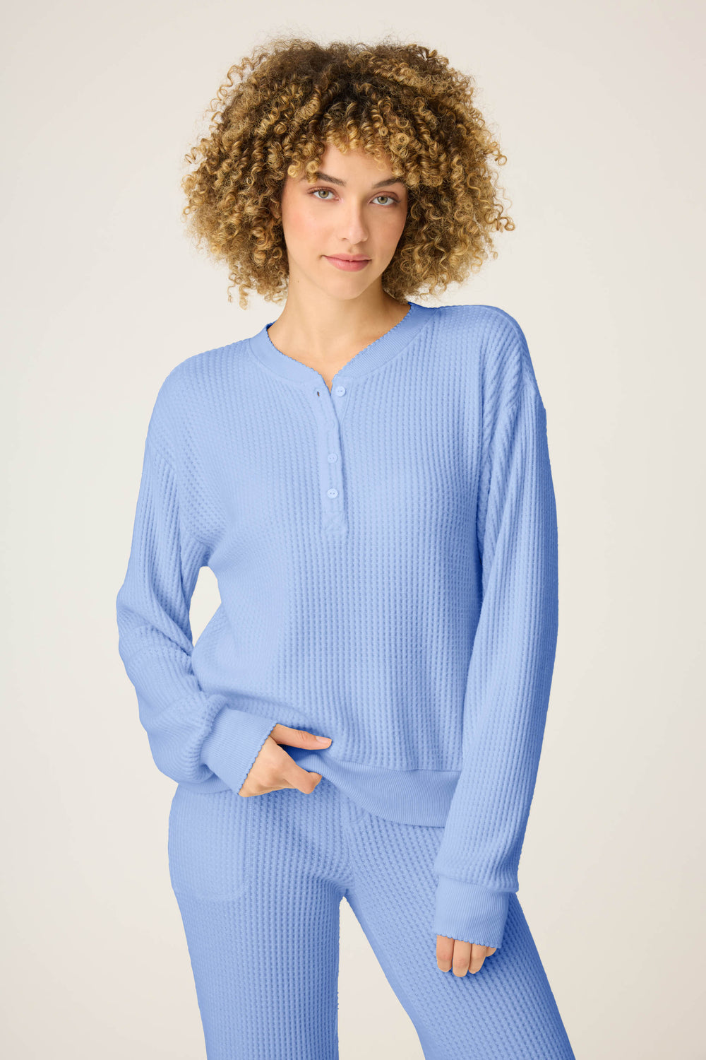 A person with curly hair wears the P.J. Salvage Morning Waffles Solid Peri Long Sleeve Top, standing against a plain light background with one hand in their pocket.