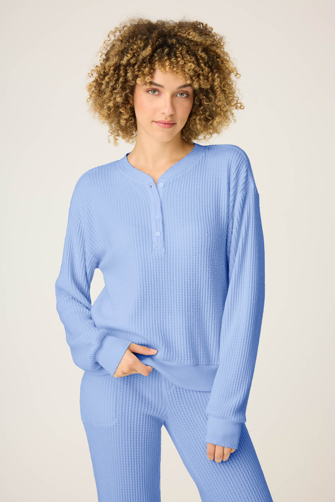 A person with curly hair wears the P.J. Salvage Morning Waffles Solid Peri Long Sleeve Top, standing against a plain light background with one hand in their pocket.
