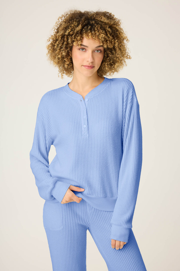 A person with curly hair wears the P.J. Salvage Morning Waffles Solid Peri Long Sleeve Top, standing against a plain light background with one hand in their pocket.