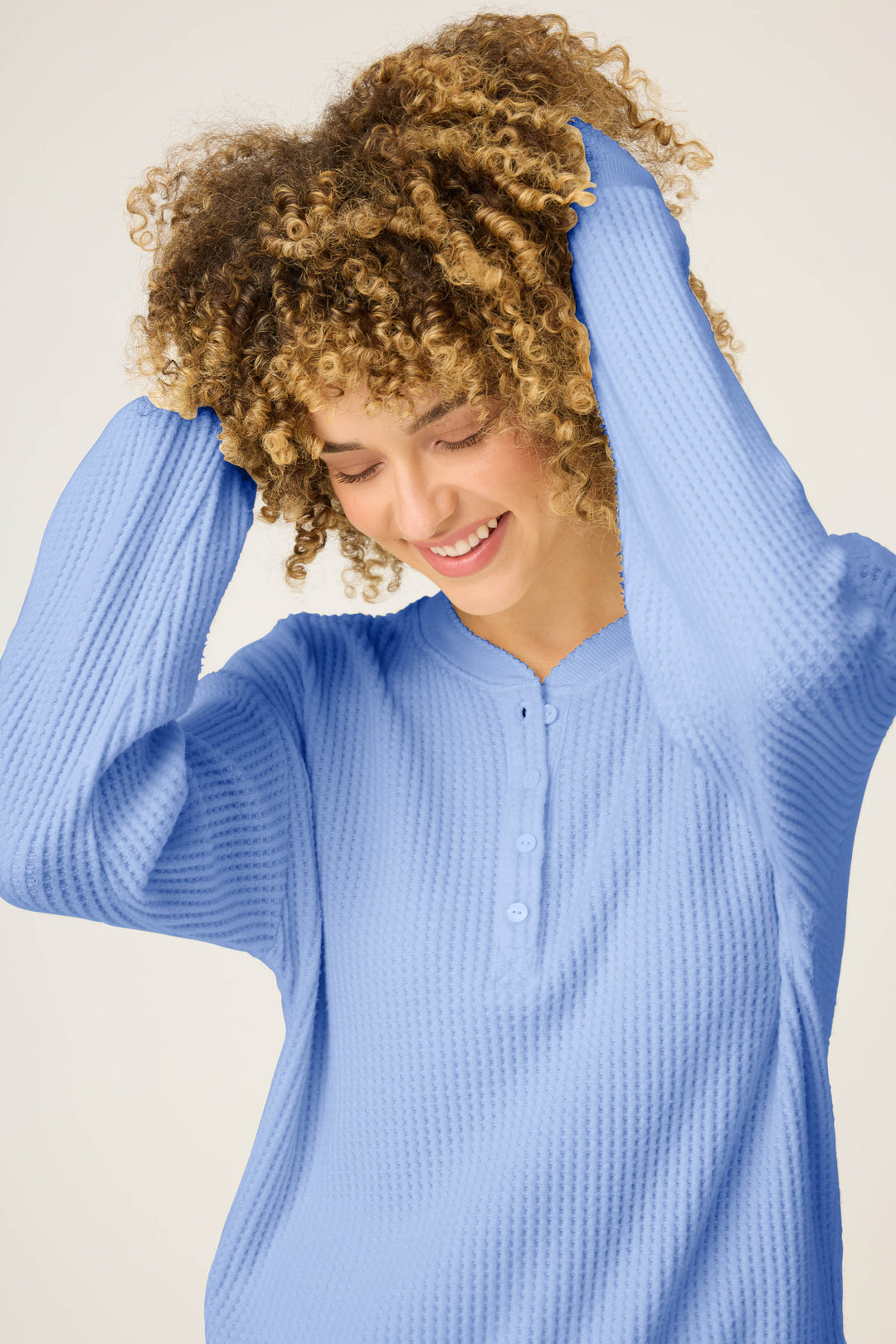 A person with curly hair smiles while looking down, holding their hair with both hands. They wear the P.J. Salvage Morning Waffles Solid Peri Long Sleeve Top. The background is plain and light-colored.