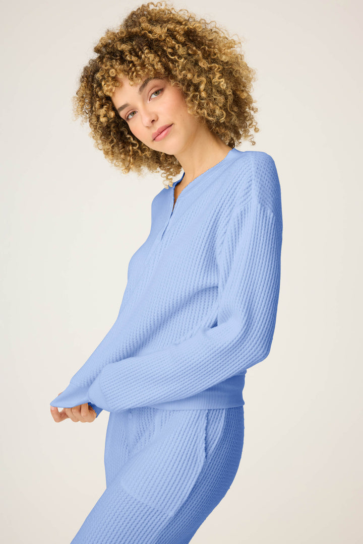 A person with curly hair wears the P.J. Salvage Morning Waffles Solid Peri Long Sleeve Top, styled with matching pants, standing against a plain light background and looking slightly toward the camera.