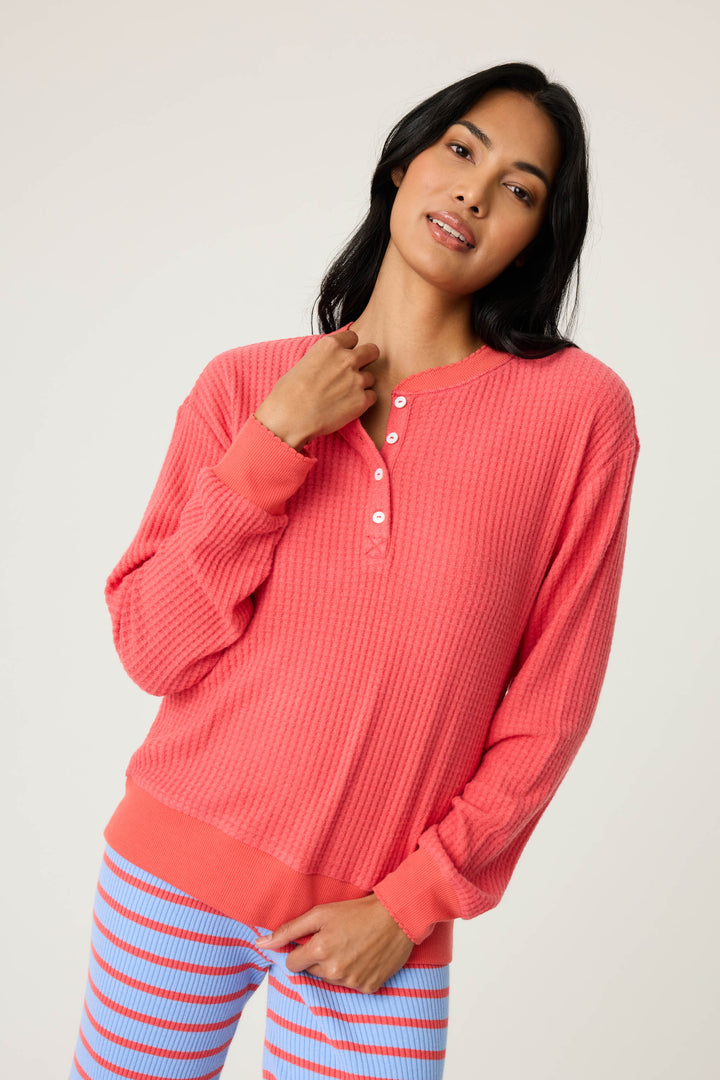 A person with long black hair wears the P.J. Salvage Morning Waffles Scarlet Long Sleeve Top, featuring a textured knit and white buttons, paired with light blue and pink striped pants against a plain light background.