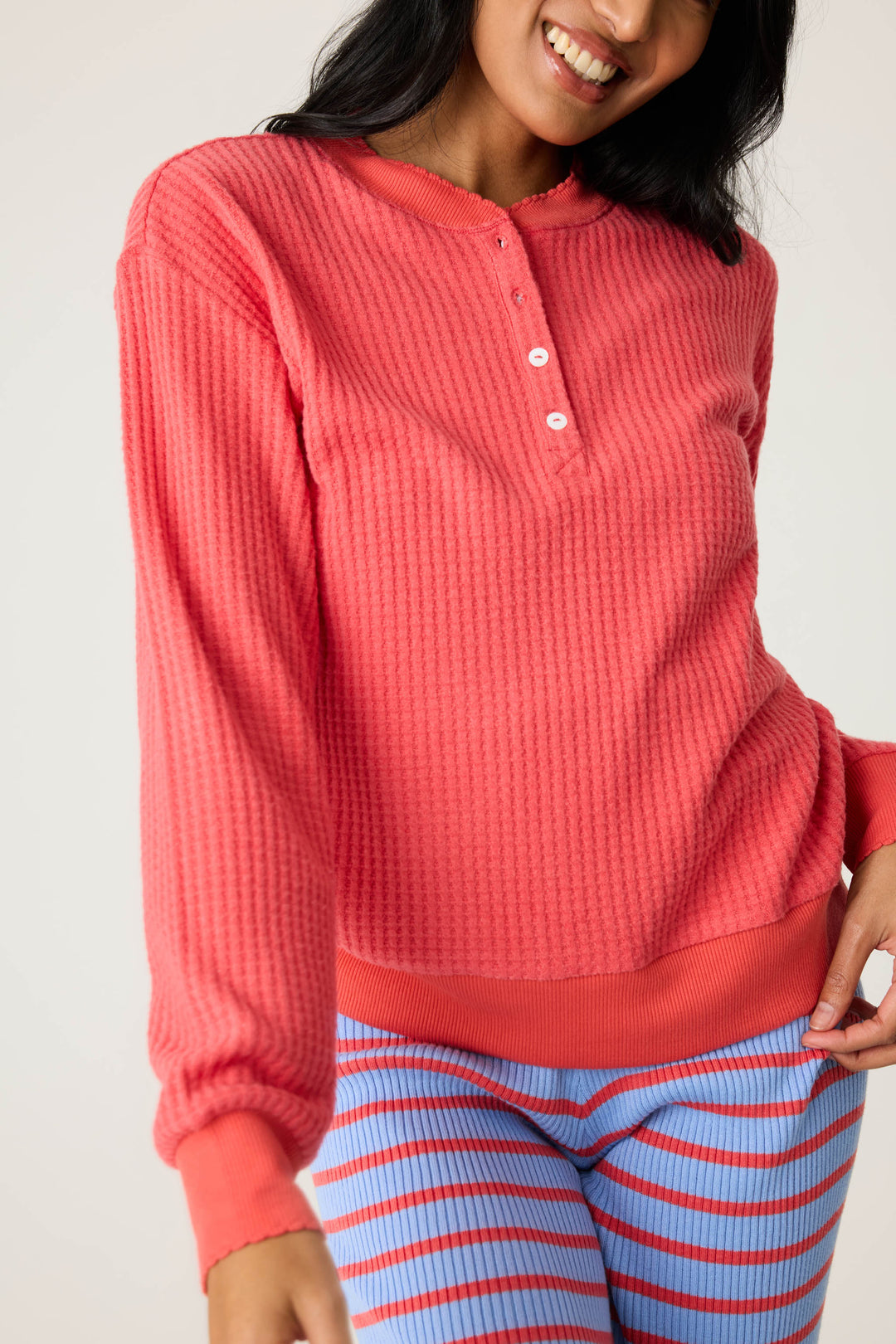 A person wears the P.J. Salvage Morning Waffles Scarlet Long Sleeve Top with blue pants featuring red stripes, standing against a plain background and smiling with one hand on their hip.