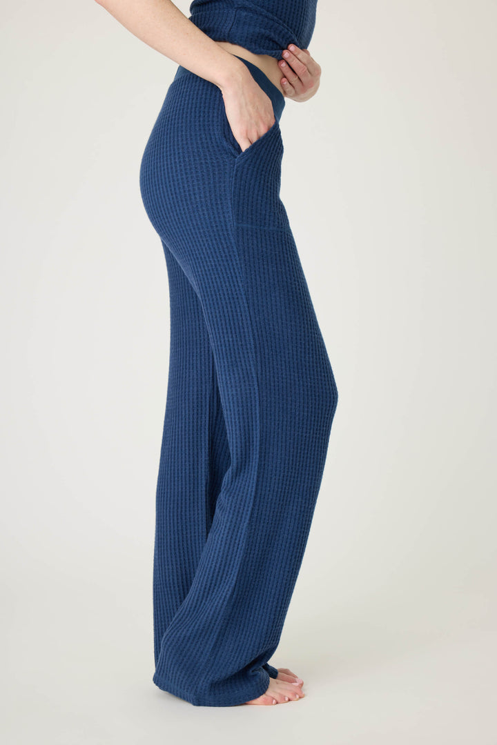 A person stands barefoot in a side view, wearing P.J. Salvage’s Morning Waffles Solid Navy Open Leg Pant with one hand in the pocket. The matching blue top complements the high-waisted, textured lounge pants against a plain light background.