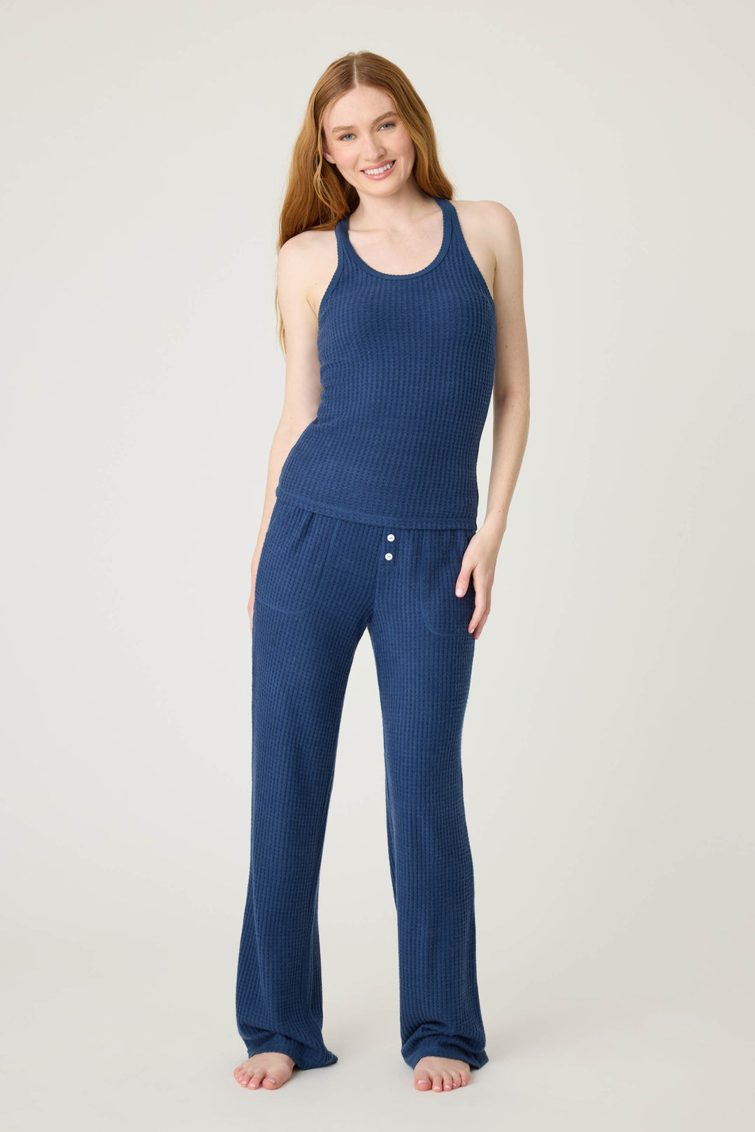 A person with long hair stands barefoot, smiling at the camera in a sleeveless blue ribbed tank top and P.J. Salvage’s Morning Waffles Solid Navy Open Leg Pant against a plain light background.