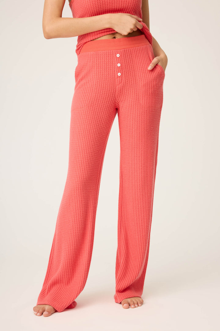 A person stands barefoot on a light background wearing P.J. Salvage's Morning Waffles Scarlet Open Leg Pant, featuring a textured look, coral color, buttons, an elastic waistband, and pockets; their hand is in one pocket with a partially visible top.