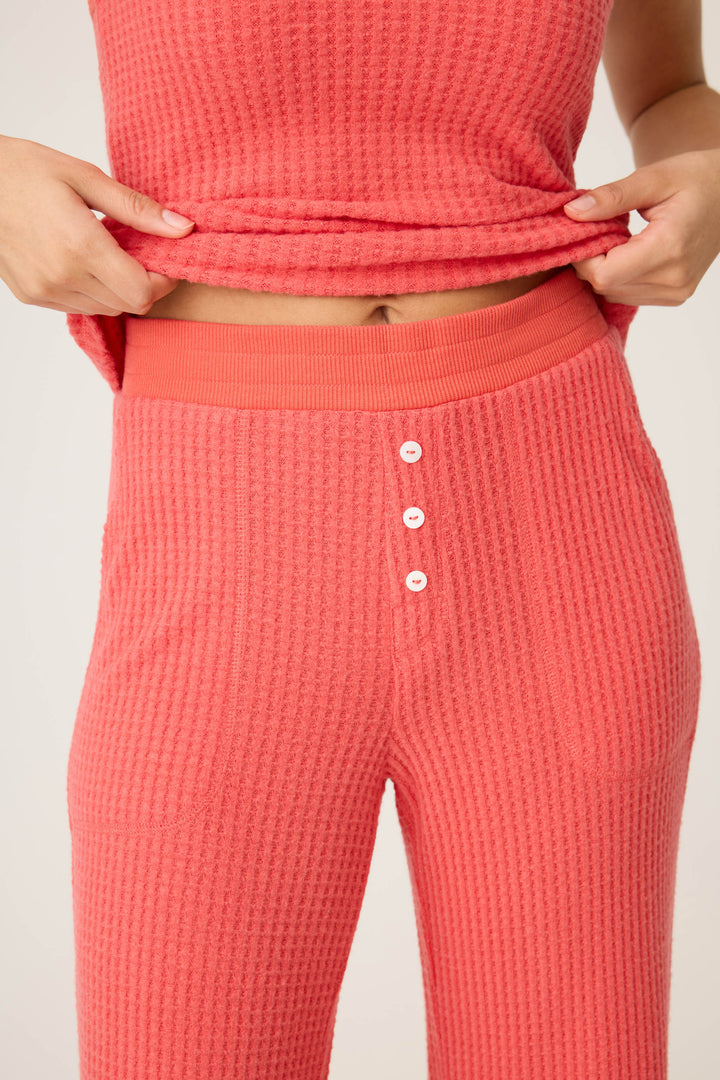 A person wearing P.J. Salvage's Morning Waffles Scarlet Open Leg Pant lifts their top slightly, showing the waistband and three white buttons on the textured coral lounge pants.