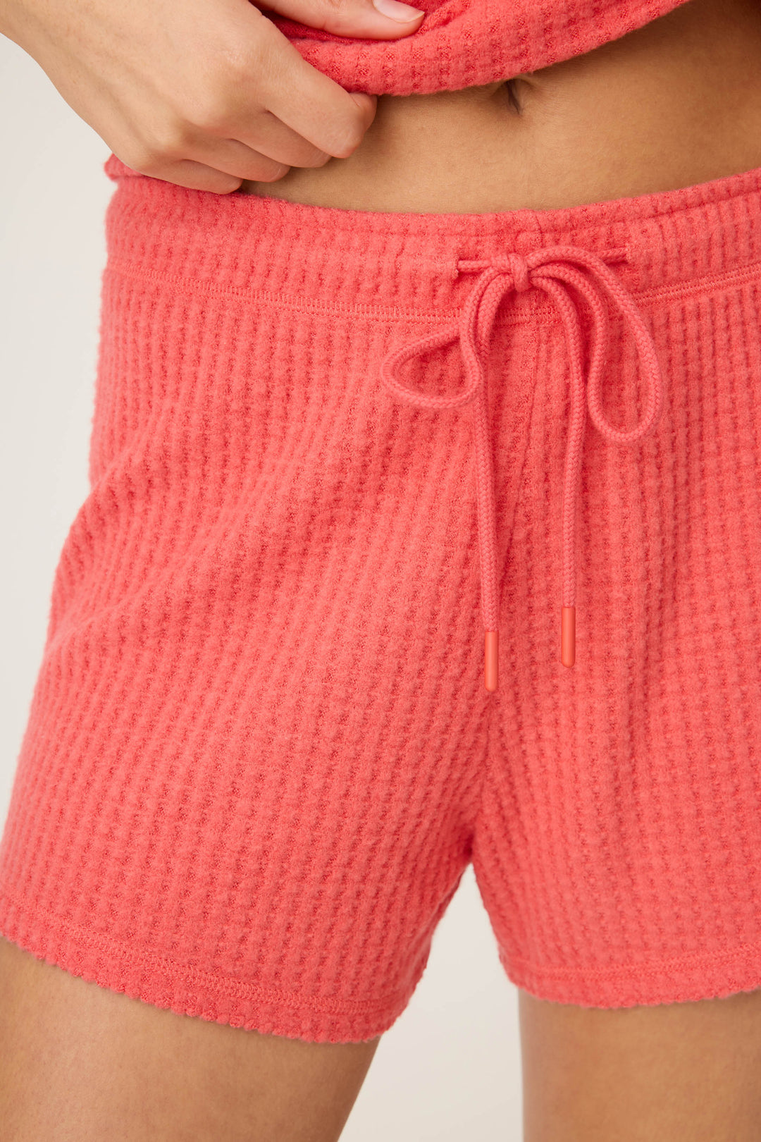 A person is wearing P.J. Salvage Morning Waffles Scarlet Shorts, featuring a textured waffle knit in coral with a front drawstring. The image shows only the lower torso and one hand.