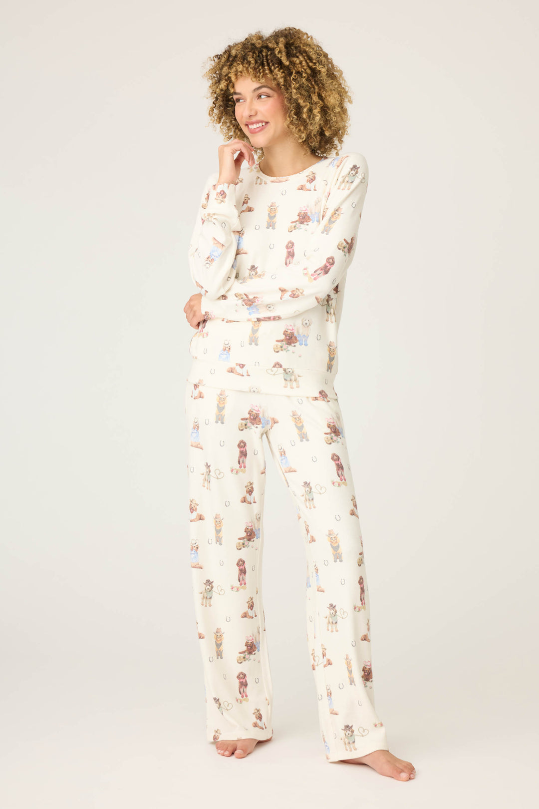 A person with curly hair stands barefoot on a light background, smiling and wearing the P.J. Salvage DOGS PJ SET RUFF ON THE RANGE—matching cream pajamas featuring a playful dog print. One hand rests on their face, the other by their side.