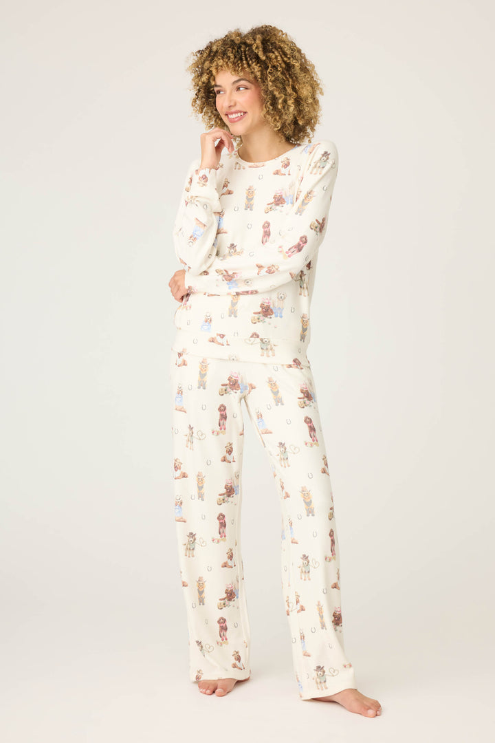 A person with curly hair stands barefoot on a light background, smiling and wearing the P.J. Salvage DOGS PJ SET RUFF ON THE RANGE—matching cream pajamas featuring a playful dog print. One hand rests on their face, the other by their side.