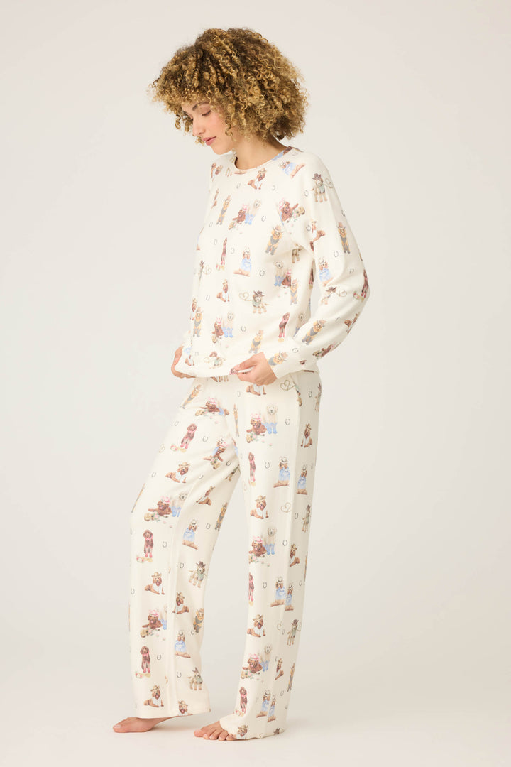 A person with curly hair stands barefoot against a plain background, wearing the P.J. Salvage DOGS PJ SET RUFF ON THE RANGE—matching long-sleeve pajama top and pants with a playful dog print—and looks down with hands in their pockets.